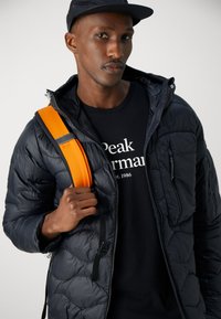 Chaqueta acolchada negra con textura de acolchado, cierre de cremallera y capucha. Lleva puesta sobre una camiseta negra. Correa de mochila de color naranja brillante cruzada sobre el cuerpo.