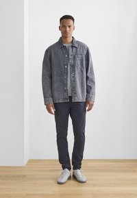 Traper jakna u izblijedjelo tamnosivoj boji, s džepom na prsima i kopčanjem na gumbe. Uparena s tamnoplavim hlačama i svijetlosivim tenisicama na drvenom podu.