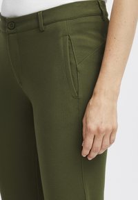 Olivnozeleni hlače iz gladkega, elastičnega materiala, ki imajo sprednji žep in prilegajočo se obliko z zankami za pas in gumbom za zapiranje.