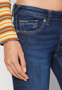 Jeans de mezclilla azul oscuro con una vista de cerca de un bolsillo frontal, con detalles de costura amarilla y roja. La mano descansa en la cadera.