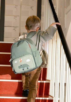 Copil cu păr castaniu deschis care poartă pantaloni maro și o jachetă verde urcă scări roșii ținând o balustradă neagră și având un rucsac verde în spate.