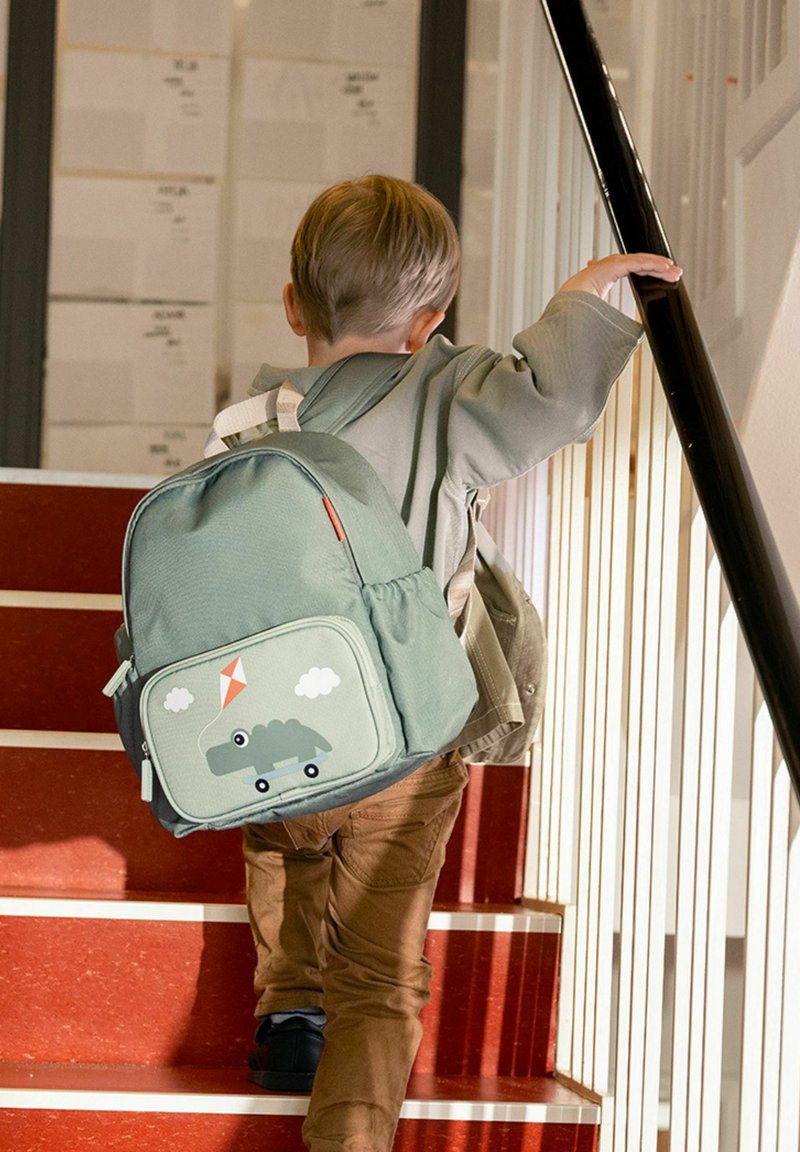 Kind mit hellbraunen Haaren, das braune Hosen und eine grüne Jacke trägt, klettert rote Treppen hinauf, hält sich an einem schwarzen Handlauf fest und trägt einen grünen Rucksack.