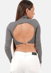 Kvinna med långt hår som bär en grå långärmad croptop med en stor cirkulär utskärning på ryggen och vita byxor med fickor.