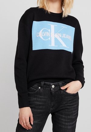Pull noir à manches longues avec un logo rectangulaire bleu portant l'inscription "Calvin Klein Jeans", associé à un jean gris foncé, main dans la poche.
