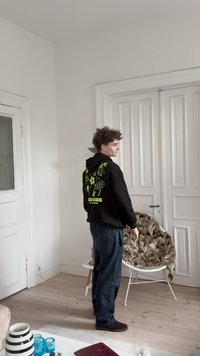 Juodas gobtuvas su neoninės žalios spalvos gėlių piešiniais ant nugaros, derinamas su laisvais mėlynais džinsais ir tamsiais batais šviesiame kambaryje.