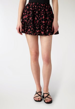 Donna che indossa una minigonna nera a fiori con piccoli fiori rossi e sandali neri con cinturini e dettagli dorati.