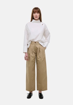 Femme debout portant un chemisier blanc à col montant, manches longues avec boutons, et un pantalon beige à jambes larges avec des chaussures noires à lacets.