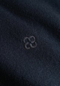 Tissu tricoté bleu marine foncé avec un petit logo géométrique brodé constitué de boucles entrelacées dans une couleur de fil assortie.