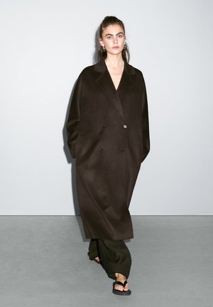 Long brown wool coat with a double-breasted design, wide lapels, and relaxed sleeves, paired with black pants and sandals on a gray backdrop.