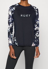 Pitkähihaiseen urheiluyläosaan kuuluu musta body, jossa on kukkakuosiset hihat. Etupuolella on valkoisella tekstillä "ROXY"-logo.