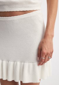 Weißer, gerippter Strickrock mit einem strukturierten Muster und einem ausgestellten, rüschengeschmückten Saum. Eine Hand mit silbernen Ringen ruht auf dem Stoff.