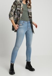 Košile s kamuflážovým vzorem, tmavě zelené upnuté tričko, světle modré úzké džíny a černé šněrovací boty. Hladké textury a přiléhavý design.