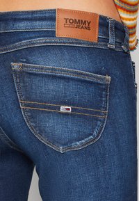 Vaqueros de mezclilla con un lavado azul oscuro, parche de cuero con la etiqueta "TOMMY JEANS" y costuras visibles en el bolsillo trasero.
