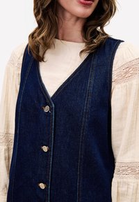Une robe salopette en denim foncé avec un décolleté en V, ornée de boutons dorés et de coutures jaunes contrastantes, portée sur un chemisier crème avec des accents en dentelle.