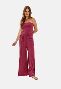 Upravený, bezramínkový overal v syté burgundské barvě, s širokými nohavicemi a nařaseným pasem. Hladká tkanina s jemnou texturou.
