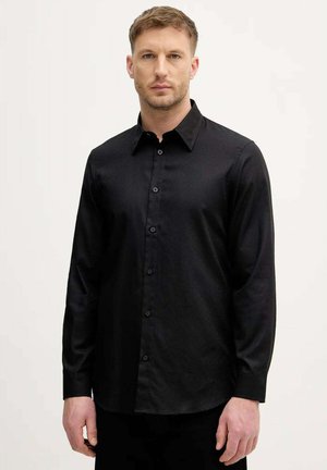 Uomo con capelli biondi corti che indossa una camicia nera a maniche lunghe abbottonata, in piedi davanti a uno sfondo chiaro e uniforme.