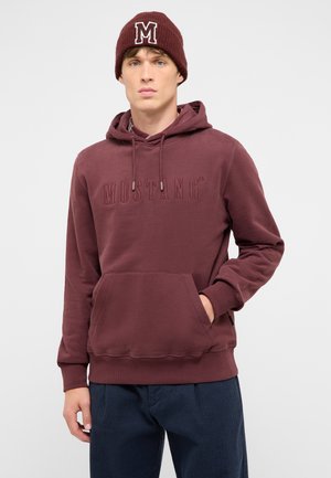 Junger Mann trägt einen bordeauxroten Hoodie mit dem Text "MUSTANG" und eine passende bordeauxrote Mütze mit dem Buchstaben M, steht mit einer Hand in der Tasche.