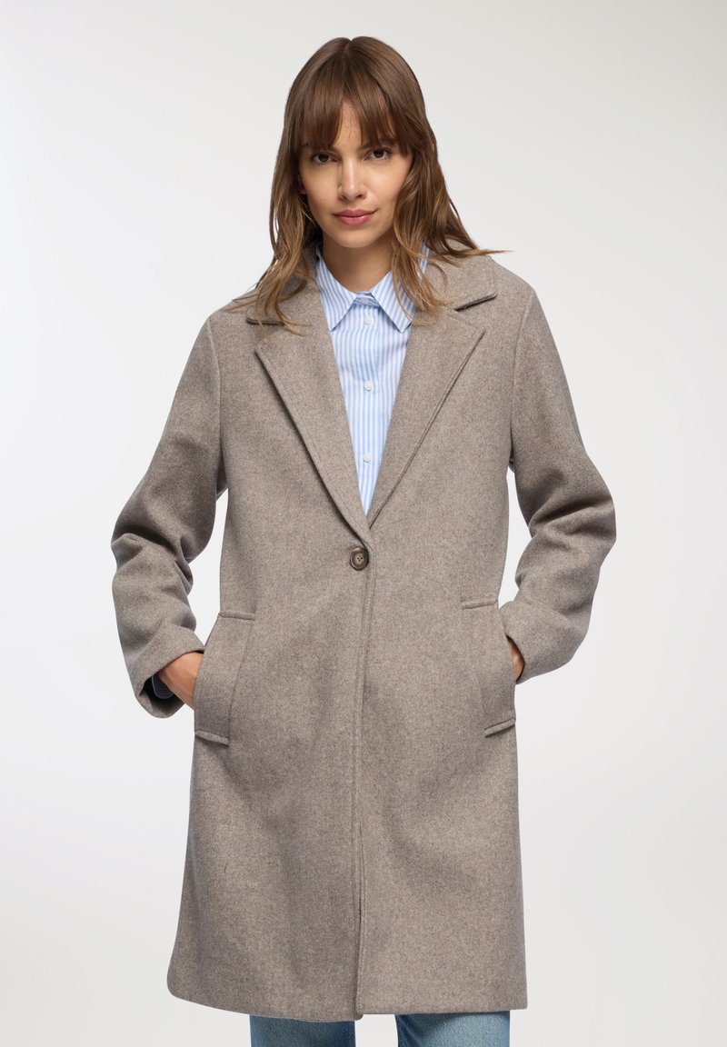 Donna che indossa un cappotto di lana beige fino al ginocchio sopra una camicia a righe blu con i bottoni, con le mani nelle tasche, su uno sfondo bianco.