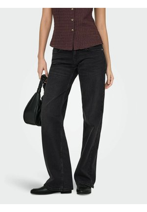 Žena oblečená v čiernych džínsach s širokými nohavicami, bezrukávovom hnedom zapínanom topi, čiernych topánkach a drží v ľavej ruke čiernu kabelku.