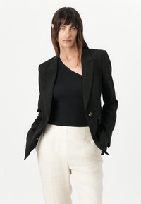 Blazer nero con vestibilità su misura e due bottoni dorati. Indossato sopra una t-shirt nera a coste, abbinato a pantaloni di lino chiari.