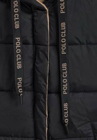 Vadderad svart jacka med dragkedja, har en beige fodring och "POLO CLUB"-märkning på svarta dragsnören. Slät textur och quiltad design.