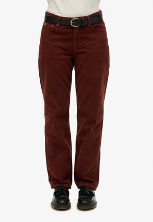 Kobieta w rdzawej sztruksowej spodniach z czarnym paskiem i czarnych skórzanych butach na jednolitym tle.