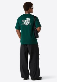 Mand iført en grøn The North Face t-shirt med logo og sorte bukser, bærende en sort skuldertaske, vendt væk mod en hvid baggrund.