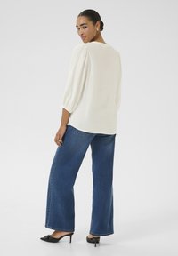 Blouse blanche texturée avec des manches bouffantes à trois quarts, associée à un jean large bleu et des chaussures à talons noires, vue de dos.