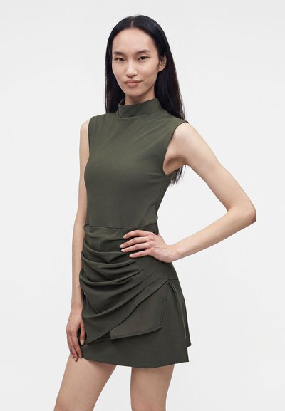 Femme debout, main sur la hanche, portant une mini-robe sans manches vert olive avec jupe froncée, face à l’avant sur un fond uni.