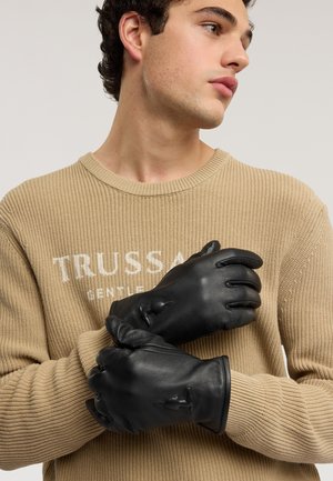 Czarne skórzane rękawiczki o gładkiej fakturze, lekko marszczone na nadgarstku, na tle beżowego, dzianego swetra z prążkowanym wzorem.