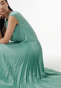 Ein plissiertes mintgrünes Kleid mit glatter Satin-Oberfläche, das ein tailliertes Oberteil und einen fließenden Rock aufweist, der elegant über den Boden fällt.