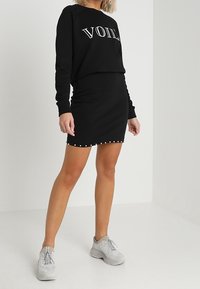 Sweatshirt noir avec impression "VOILÀ", associé à une jupe noire ajustée comportant des clous blancs le long de l'ourlet. Le modèle porte des baskets gris clair.