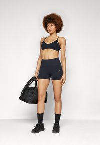 Soutien-gorge de sport noir et shorts taille haute en matériau extensible. Le modèle tient un sac de sport noir. Des chaussettes noires et des chaussures de sport complètent le look.