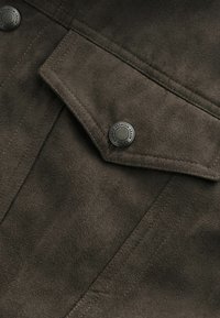 Poche de veste en daim brun foncé avec rabat pointu sécurisé par un bouton-pression métallique rond gravé de "Premium Grade Durable Goods."