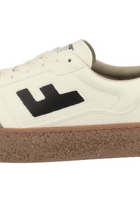 Valkoinen nahkainen lenkkari, jossa on musta logo, beige korostukset ja teksturoitu ruskea kumipohja. Sisältää klassisen nauhakiinnityksen ja sileän pinnan.