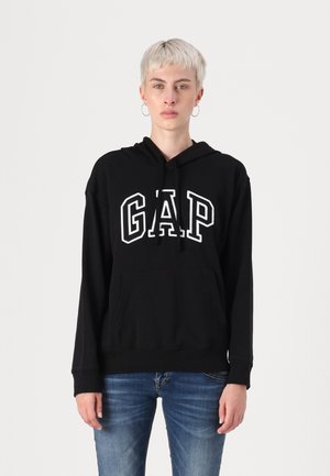 Svart hettegenser med hvit "GAP"-logo, frontlomme som en kenguru, lange ermer og ribbestrikket kant. Båret med blå jeans.