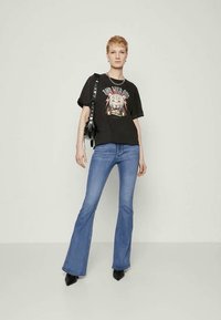 Črna grafična majica s dizajnom leva, v kombinaciji z modrimi širokimi jeans hlačami in črnimi čevlji na peto. Model drži črno torbico.