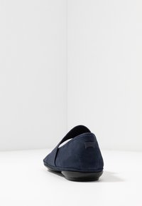 Chaussure plate en daim bleu marine avec des brides croisées élastiques et une semelle noire, vue de dos sur une surface blanche.
