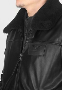 Veste en cuir noir avec un col en shearling, fermeture éclair et un patch texturé avec un logo d'aigle. Texture douce, design ajusté.