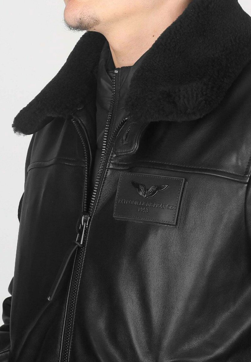 Veste en cuir noir avec un col en shearling, fermeture éclair et un patch texturé avec un logo d'aigle. Texture douce, design ajusté.