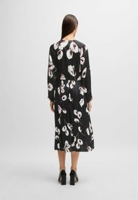 Schwarz Kleid mit langen Ärmeln, florales Muster in weiß und rot, Taillendetails und mittellange Länge. Getragen mit schwarzen Blockabsatzschuhen.