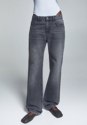 Personne portant un jean noir délavé et ample associé à des ballerines noires à bout carré sur un fond gris uni.