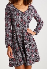 Abito floreale in bianco e nero con maniche lunghe, scollo a V e gonna svasata; presenta dettagli rossi e un tessuto leggero e testurizzato.