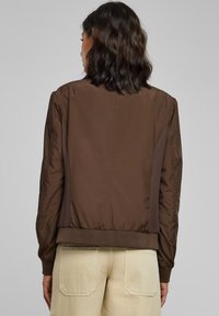 Giacca bomber marrone con materiale liscio e lucido, caratterizzata da polsini e orlo a costine, dettagli laterali in rete e una vestibilità morbida.