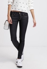 Jeans negros ajustados con un diseño de cinco bolsillos, costuras en contraste y una textura suave, combinados con un suéter blanco de lunares y zapatillas.