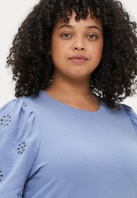 Ανοιχτό μπλε πούλοβερ με στρογγυλή λαιμόκοψη και φουσκωτά μανίκια, διακοσμημένο με λουλουδάτα σχέδια από ματόχαντρα στους ώμους, κατασκευασμένο από μαλακό βαμβακερό ύφασμα.