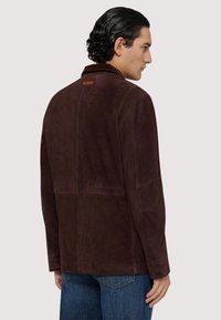 Brun mockaläderjacka med krage, långa ärmar och en detalj bak. Har en slät textur och en strukturerad silhuett.