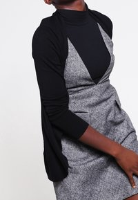 Cardigan nero a maniche lunghe sopra un dolcevita nero a coste, abbinato a un vestito grigio testurizzato con scollatura a V e tasche.