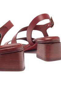 Sandalias de tacón de cuero rojo con tacón cuadrado, que presentan correas entrecruzadas y un detalle de hebilla metálica circular.
