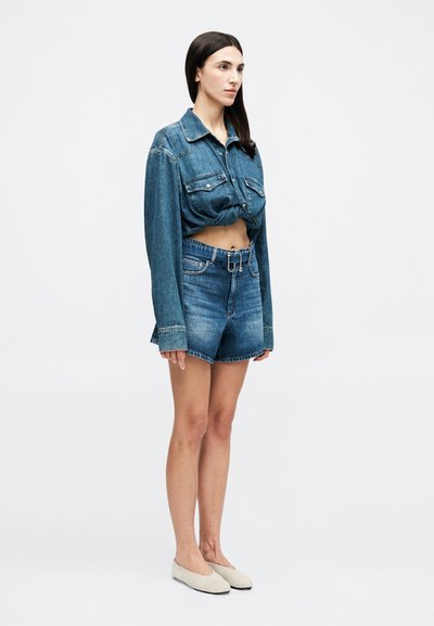 Γυναίκα με μακριά σκούρα μαλλιά, που φορά cropped τζιν μπουφάν, σορτς τζιν με ψηλή μέση και ζώνη, και μπεζ επίπεδα παπούτσια, στέκεται πλαγίως.